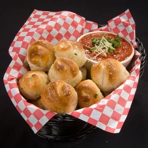 Angelo's Pizza Parlor garlic knots with marinara sauce, garnished with parsley and cheese, in a basket.