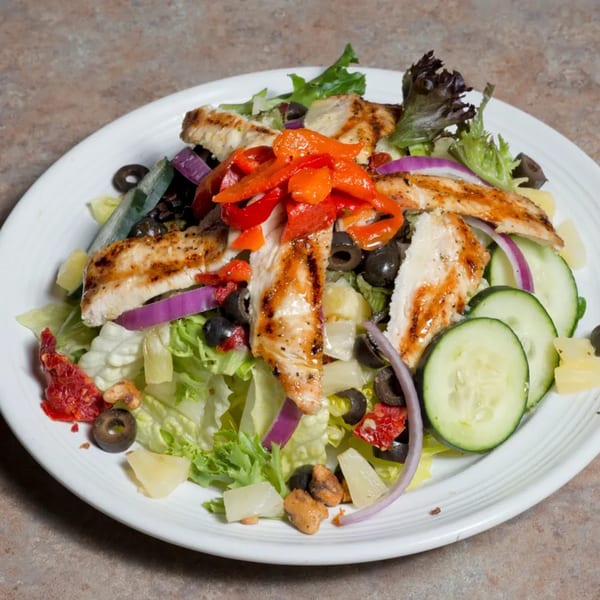 Grilled chicken salad with lettuce, red peppers, olives, cucumbers, and red onion on a white plate.