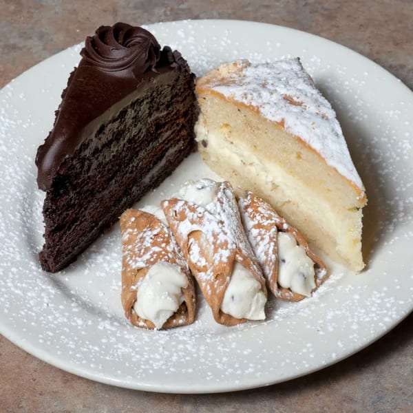 A white plate holds a slice of chocolate cake, a slice of cheesecake, and three cannolis.